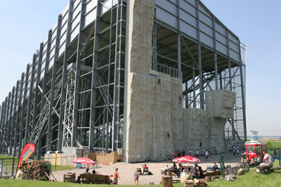 Große Kletterwand im Freien, an der Menschen klettern und andere auf Bänken und an Tischen darunter sitzen, umgeben von einem Metallgerüst und einem offenen Himmel.