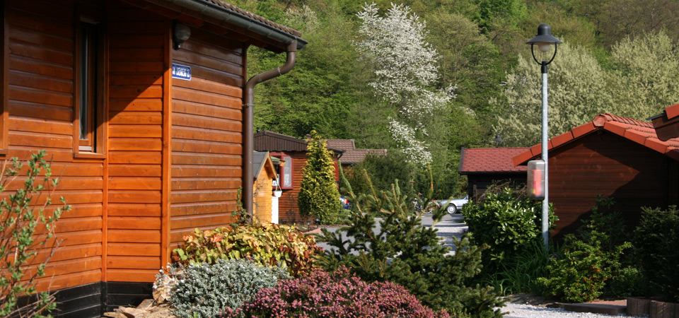 Die Holzhütten sind entlang eines Weges mit Sträuchern und Laternenpfählen aufgereiht, umgeben von Grün und Bäumen in einer hügeligen Gegend.