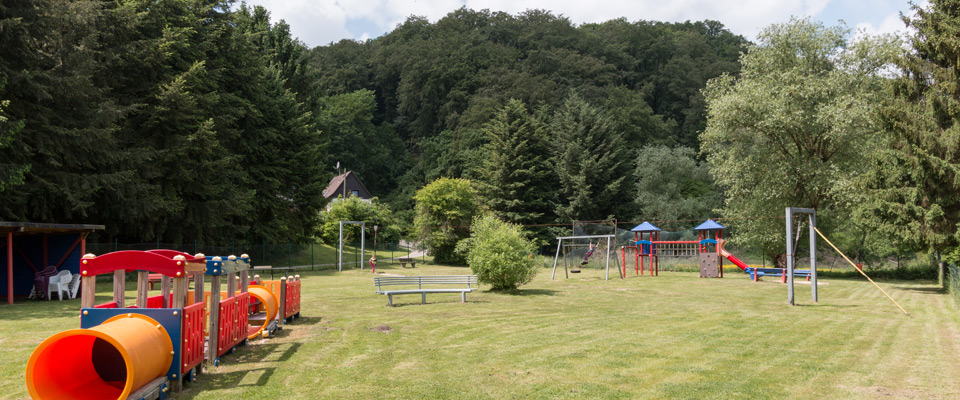 Spielplatz im Freien mit Rutschen, Klettergerüsten, Bänken und einer Rasenfläche im Freizeitdomizil Ruhrtal, umgeben von Bäumen und bewaldeten Hügeln im Hintergrund.