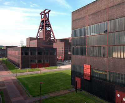 Ein Komplex großer Industriegebäude aus rotem Backstein mit einem hohen Förderturm aus Metall und grünen Rasenflächen unter einem teilweise bewölkten Himmel.