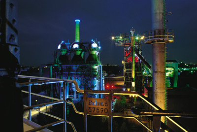 Industrieanlage, die nachts mit bunten Lichtern beleuchtet wird, mit Metallstrukturen, Rohren und einem Schild mit der Aufschrift Behm & 57590 im Vordergrund.
